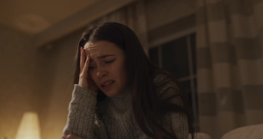 A woman sitting in a room crying with a window and a lamp in the background