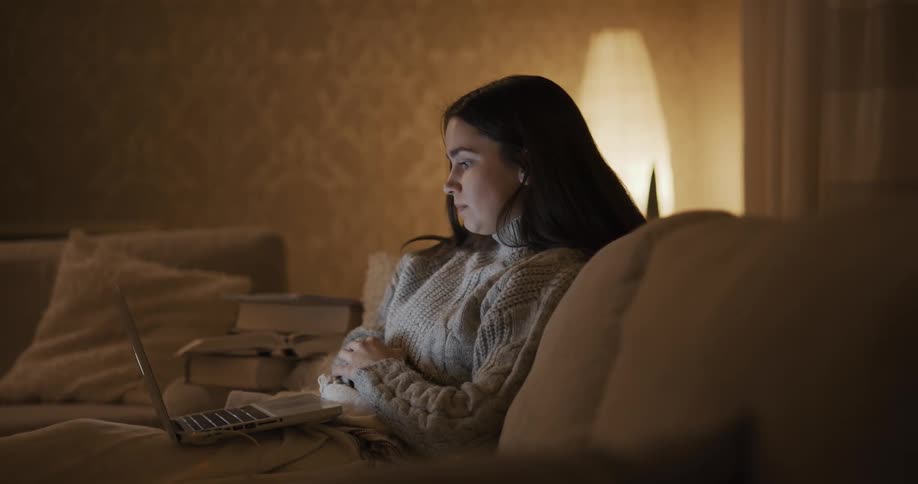 A young woman sitting under a blanket on a sofa in the living room of a house participates in a video chat using the laptop sitting in her laptop