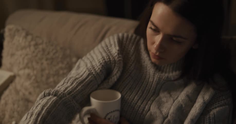 A woman sitting on a couch holding a mug reading a book and writing notes