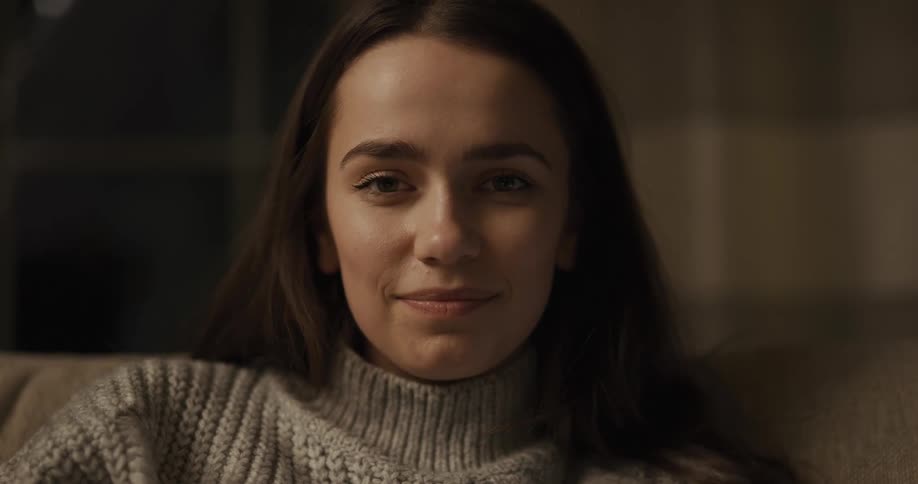 A young woman in a sweater sitting on a sofa smiles into the camera
