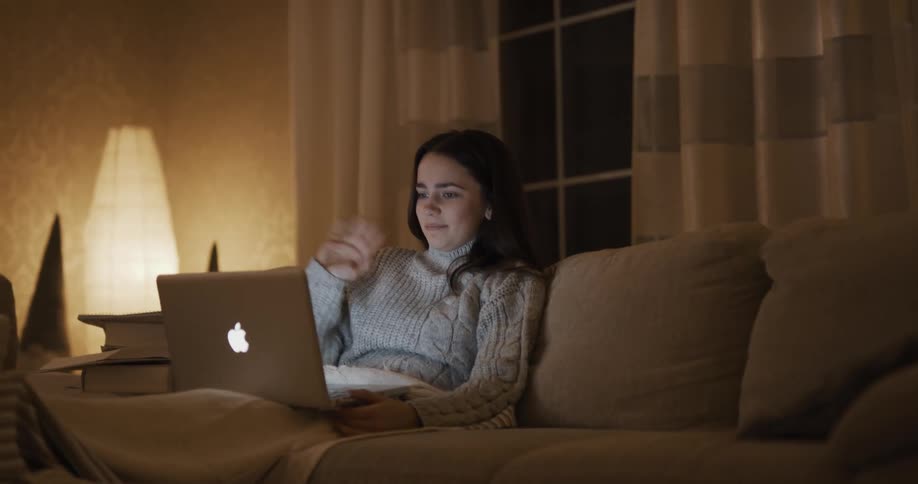 A woman sits on her couch during a video call on her laptop and waves goodbye