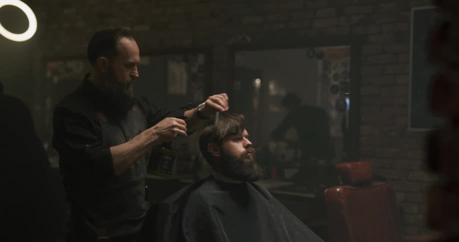 A man in a barbershop spraying and combing a mans hair with a wall and a mirror in the background