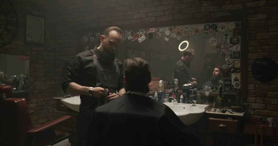 A barber puts on a black apron and talks to a client in the salon chair