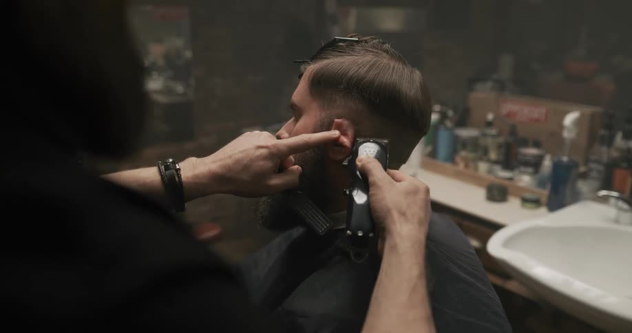 A man sits in a barber chair while another man uses clippers to trim his hair