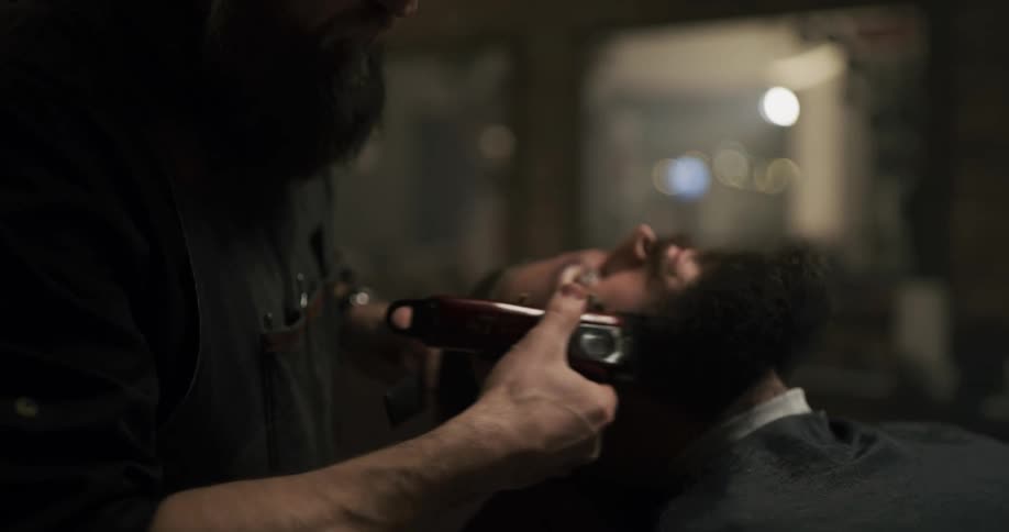 A barber with a full hipster beard trims and combs the beard of a customer in his chair