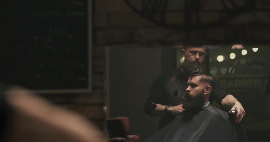 A man in a barbershop sitting on a chair getting a haircut with a wall and a mirror in the background