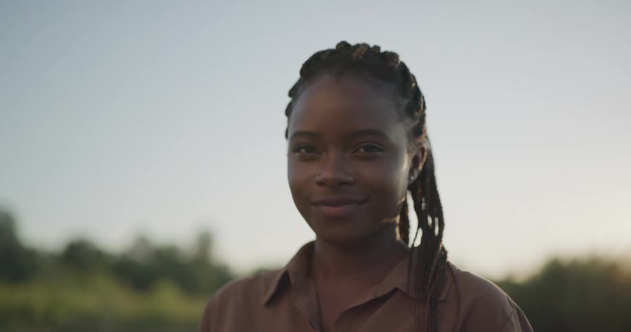 A young black woman smiles into the camera while standing outdoors in a field during the day