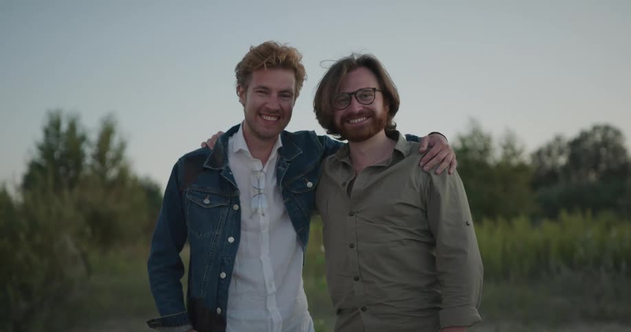 Two men stand side by side with arms around each others shoulders as they smile