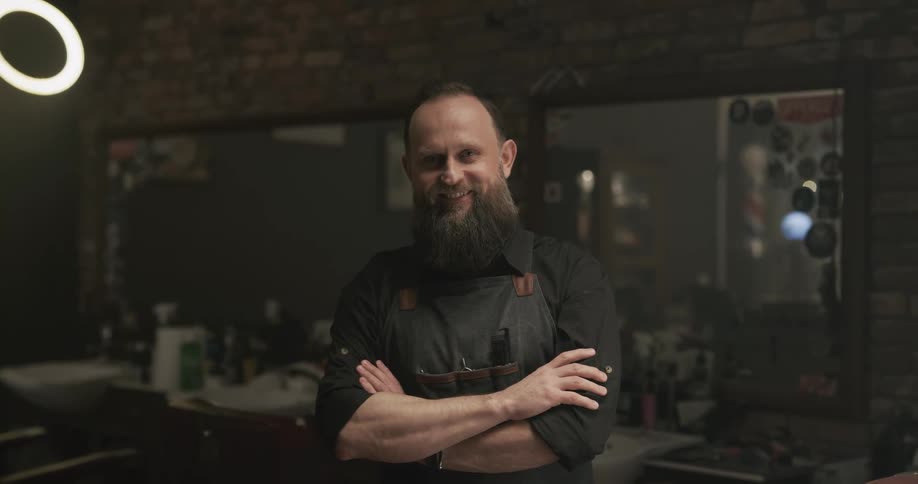 Male barber with black shirt apron and beard faces the camera with eye contact and smiles