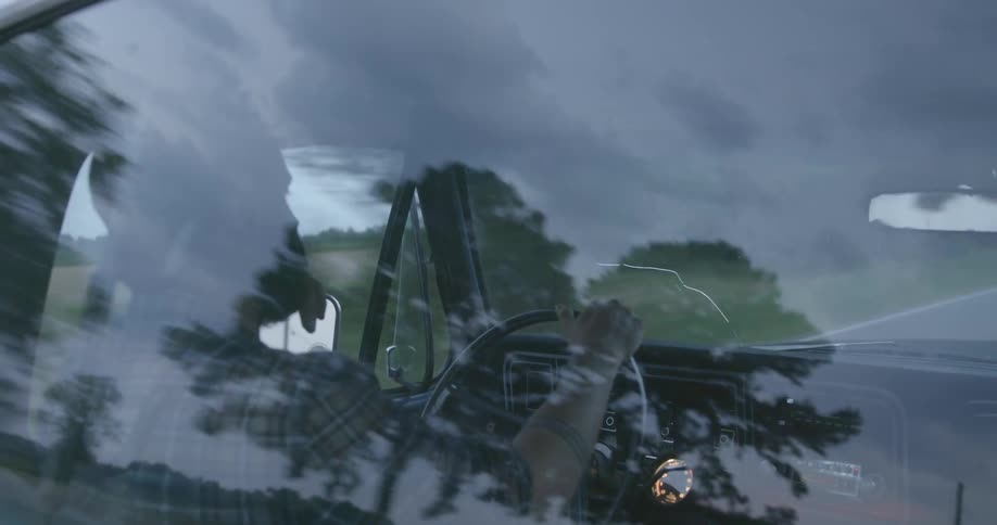 A man with a hat driving a vehicle with trees the sky and clouds in the background and reflecting off the window