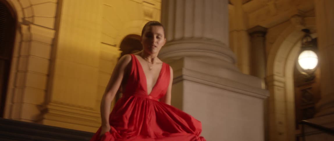 A woman in a long red evening dress rushes down stairs outside of a large building
