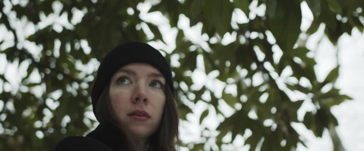 A woman wears a black coat and knit hat while standing in front of a large tree