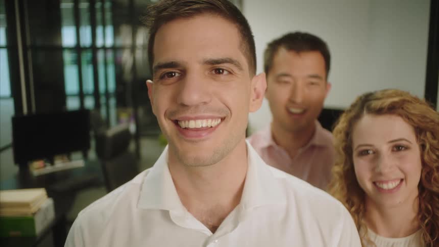 A man pats another man on the back as they stand in an office and talk with someone