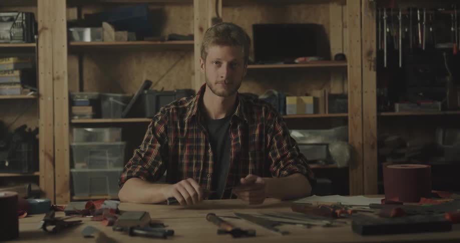 A man poses behind a table in his workshop holding a knife as other tools surround him