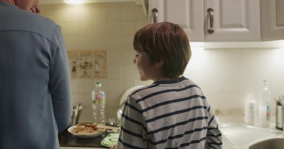 A boy running into a kitchen with a woman cooking pancakes