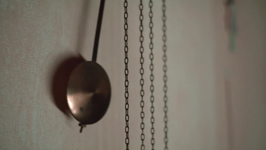 Cuckoo clock pendulum bob swinging behind the winding chains over a wall