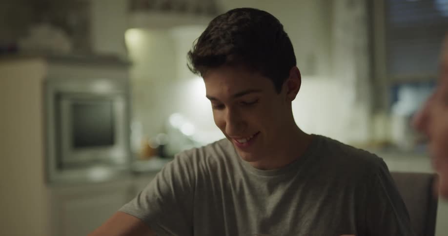 A woman sitting at a dining table in a home kitchen hands a plate of berries to a young man who uses his fork to push some of the berries onto his plate