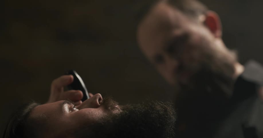 Barber at a barber shop trimming the beard of his customer with a beard trimmer