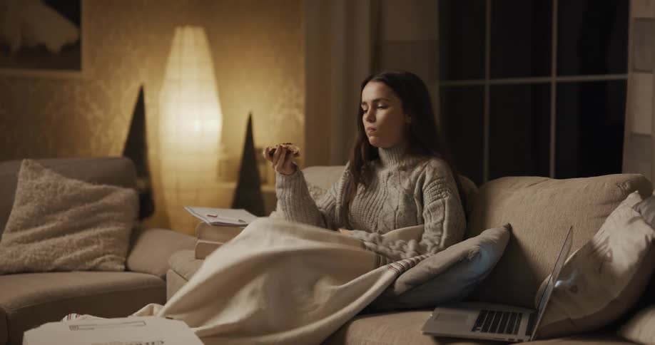 A female carries a pizza box and sits down on the couch to start eating