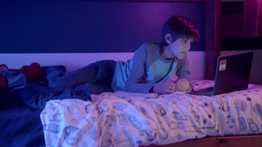 A boy laying in a bed writing in a book with a pencil in front of a laptop