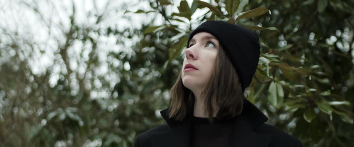 Woman wearing black knit hat and a black jacket is looking around in an area with a lot of trees