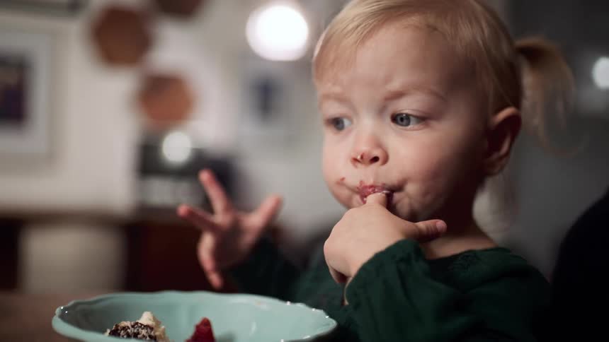 A little girl in a room sitting on a chair at a table eating dessert