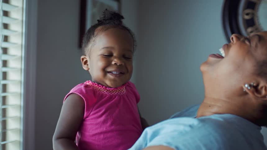 A woman in a room holding a little girl next to a window with a wall and a clock in the background