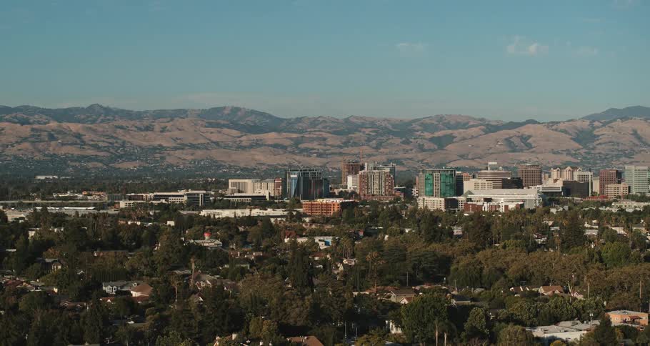 A city with buildings with trees in the foreground with a mountain range the sky and clouds in the background