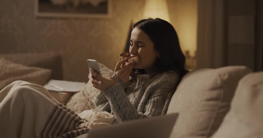 Female sits on the couch while scrolling on her smart phone and taking a bite of pizza