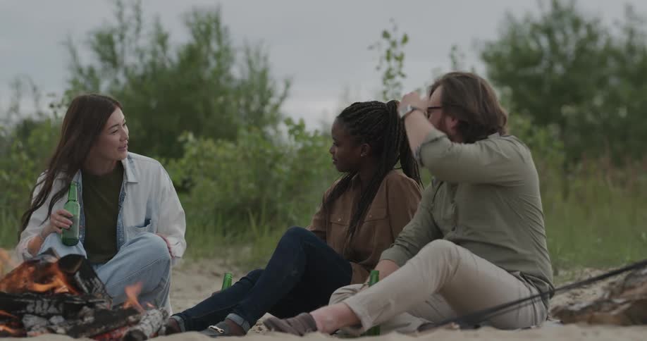Group of friends sitting together around a bonfire while chatting and drinking beverages