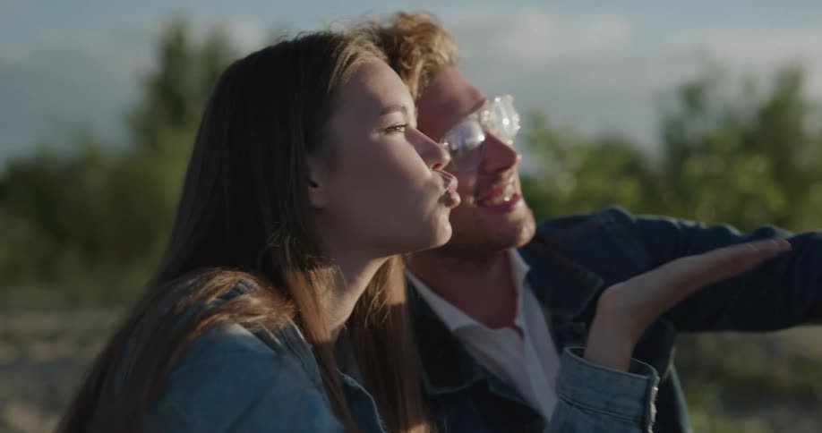 A man and woman sitting on the beach take a selfie with a cell phone