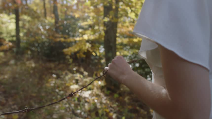 A woman standing in a forest touching barbwire with trees the sky and clouds in the background