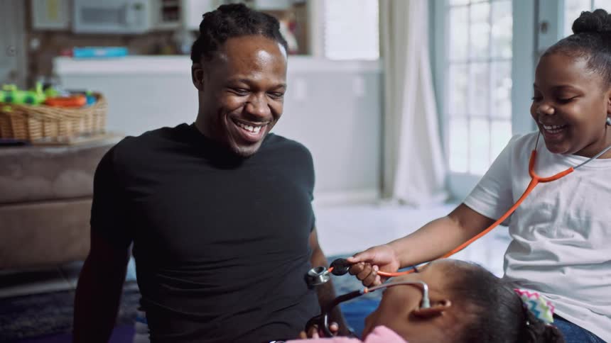 A laughing man sits on a rug in the living room of a house as his daughters examine him with a pair of stethoscopes