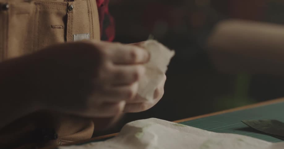 A man sits at a table in a workshop apron and buffs a sample of leather with a cloth