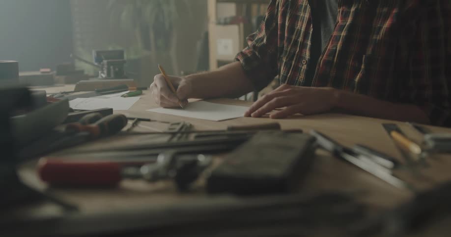 A man draws lines with a pencil on a piece of paper as he sits at a desk littered with small tools