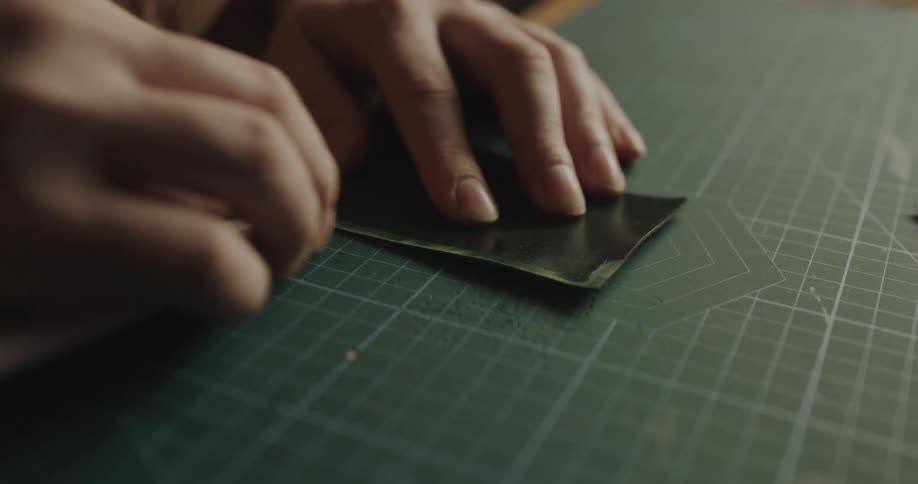 A man uses a small blade to scrape the edges of a black sheet of paper on a green gridded tabletop