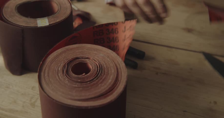 Hands tear off a piece of abrasive cloth from a roll sitting on a workbench with other tools