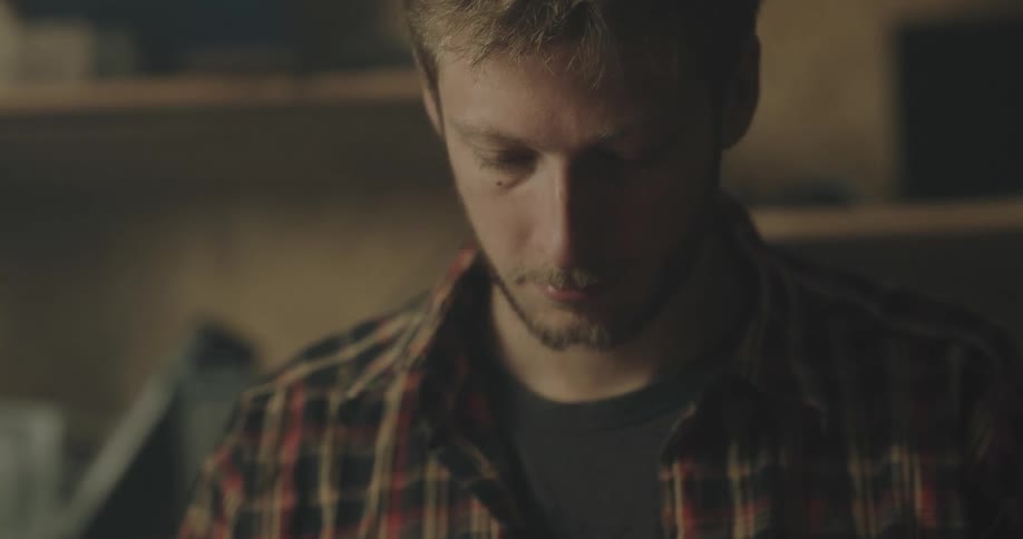 A man wearing a plaid shirt over a tee looks down as he works