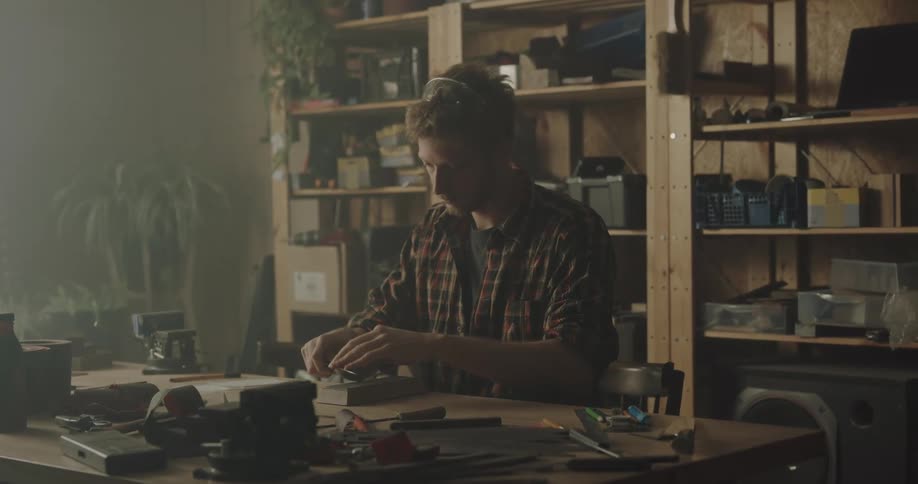 A man sitting on a chair at a table sharpening a knife with shelves in the background