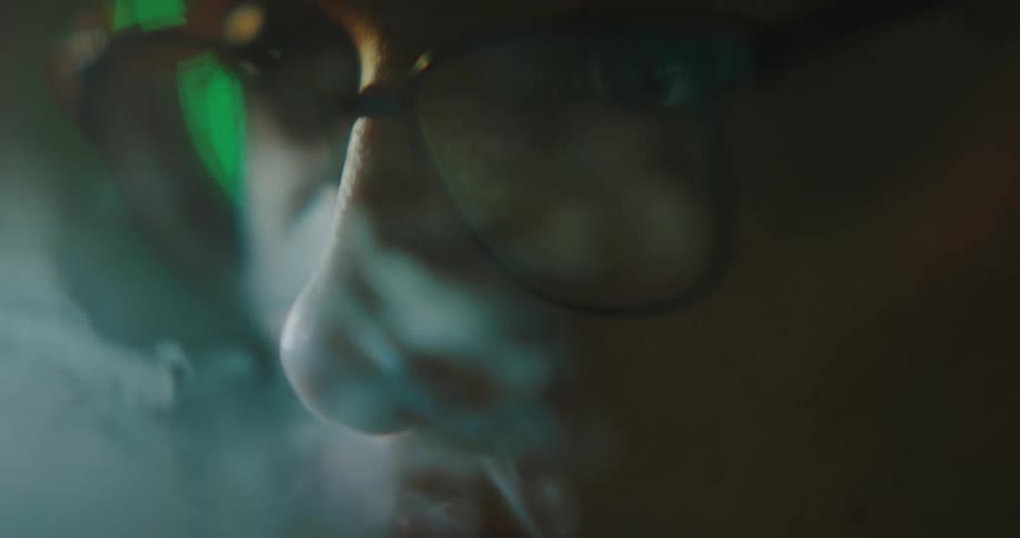 White man wearing glasses and a hat smoking while using a computer