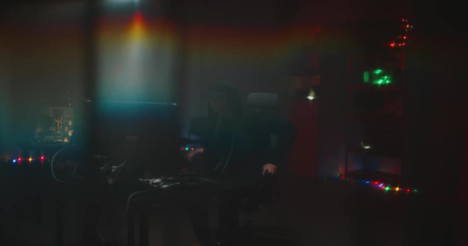A man sits down at a desk in front of two computer monitors in a server room with multiple colorful and flashing lights