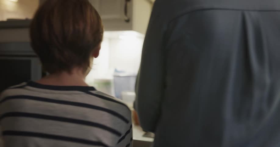 A boy runs into the kitchen to examine the food being prepared by a woman before the woman shoos him away