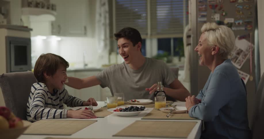 A woman and two boys sitting at a table with a refrigerator and windows in the background