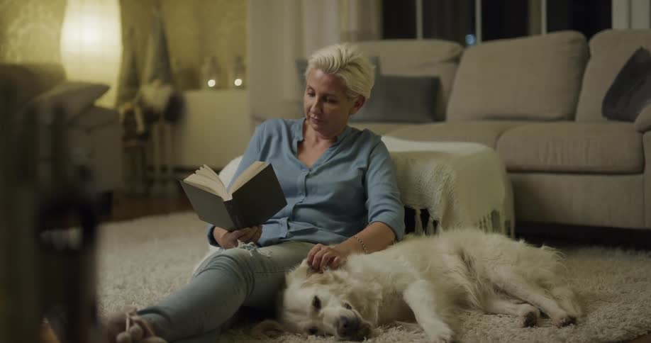 A woman is sitting and reading a book while she pets her dog