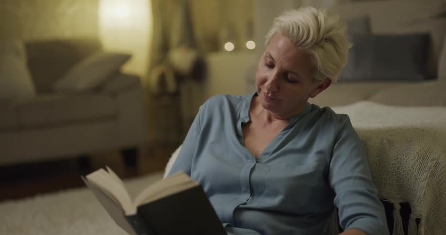 White woman sitting at a rug and petting her dog while reading a book