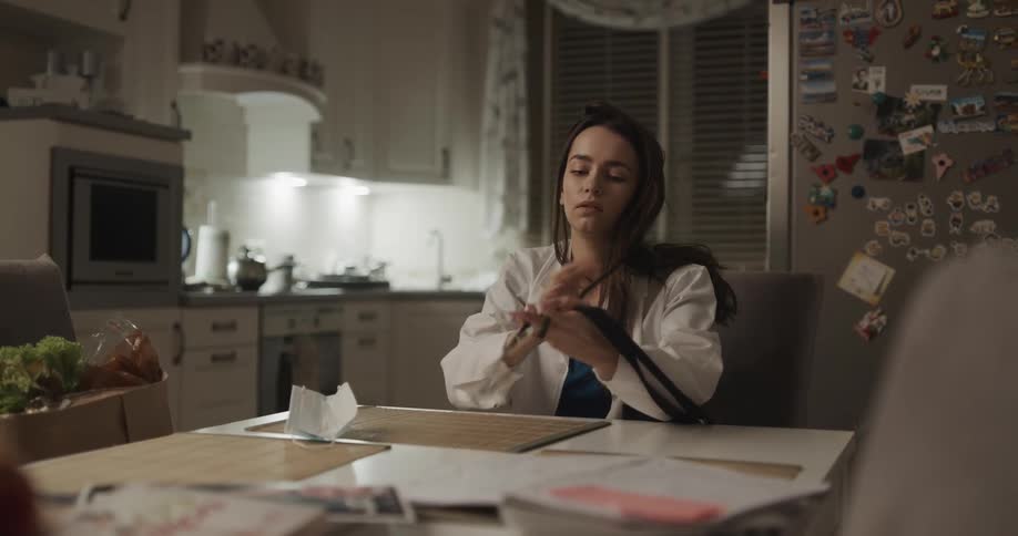 A doctor walks into her kitchen carrying a paper bag of groceries and sits down