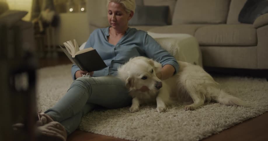 A woman in a room sitting on the floor reading a book and petting a dog