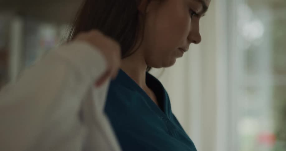 A woman in a room putting on a lab coat and a stethoscope around her neck with a window in the background