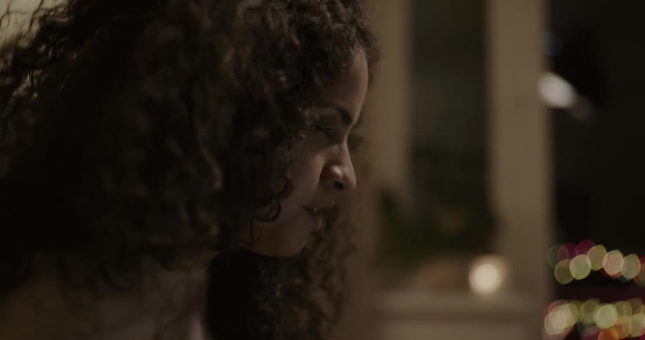 A woman with dark curly hair speaks animatedly in front of a window at home