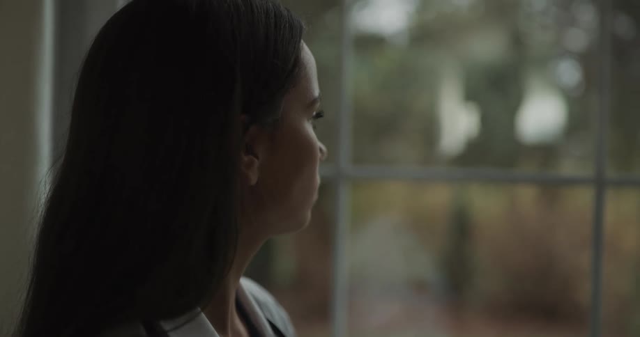 Female doctor looking through a window in a contemplative way and then putting on a mask